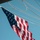 American Flag USS Gladiator (MCM 11) A large American flag is captured mid-wave against a clear, cloudless blue sky, photographed from a low angle looking upward. The flag appears to be mounted on a vessel or structure, as multiple natural-fiber ropes and rigging lines are visible in the background. The image is sharp and well-lit, suggesting bright daylight conditions.