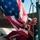 American Flag USS Gladiator (MCM 11) A person is handling a large American flag aboard what appears to be a naval or maritime vessel. The individual is gathering or securing the flag near rigging, ropes, and metal railings on the ship's deck. The scene is captured in bright daylight with the ocean visible in the background.