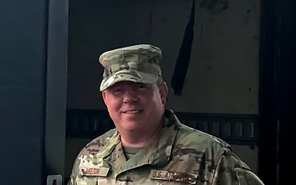 A male service member in U.S. Air Force Operational Camouflage Pattern (OCP) uniform is posing with hands on hips and smiling. He is wearing a matching OCP patrol cap and has visible name tape and 'U.S. AIR FORCE' tape on his uniform. A sign reading 'ANTHONY' is visible on the wall behind him.