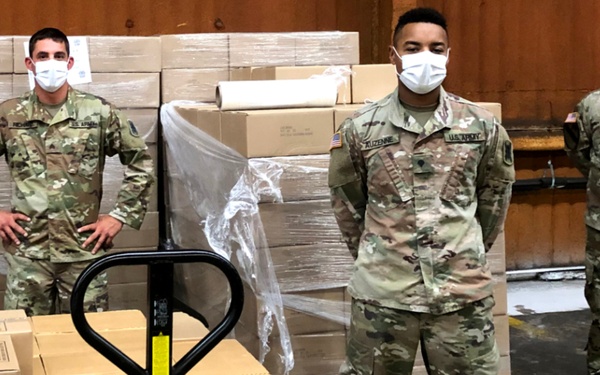 Three U.S. Army soldiers in MultiCam camouflage uniforms and white face masks are standing in a warehouse surrounded by stacked cardboard boxes on pallets. A manual pallet jack loaded with boxes is visible in the foreground. This appears to be a logistics or supply distribution operation, likely during the COVID-19 pandemic given the masks and warehouse setting.