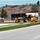 A yellow John Deere motor grader (model 670G) is actively grading or leveling soil near a roadway at what appears to be a construction or development site. The scene includes newly laid pavement, fresh sod, storage containers, and fencing in the background. The setting appears to be a semi-rural area with a hillside visible in the background.