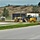 A yellow John Deere motor grader is parked or operating near a road median in what appears to be a light industrial or commercial area. The scene shows a well-maintained roadway with grass medians, chain-link fencing, storage containers, and a hillside with mixed vegetation in the background. The setting suggests active road construction or grading work in progress.
