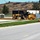 A yellow John Deere motor grader (model 670G) is actively grading or leveling soil along a roadside construction site. The scene includes a paved road in the foreground, freshly laid sod or grass strips, and several storage containers in the background. The setting appears to be a commercial or industrial development area with a hillside visible in the distance.