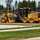 A John Deere 670GP motor grader is actively grading a dirt/gravel surface at a construction site. The yellow heavy equipment bears the model number '670GP' and 'DEERE' branding prominently on its body. The site is bordered by a chain-link fence, stacked pipes, and evergreen trees in the background.