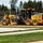 A John Deere 670GP motor grader is actively grading a dirt surface at what appears to be a construction or road preparation site. The yellow heavy equipment is positioned on exposed orange-brown soil, with a paved road and green grass visible in the foreground. A fenced storage yard with stacked pipes or cylinders is visible in the background.