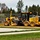 A John Deere 670GP motor grader is actively grading a dirt/gravel surface at what appears to be a construction or infrastructure site. The yellow heavy equipment is positioned between a paved road and a fenced storage yard containing stacked pipes or culverts. The setting includes a mix of grass, gravel, trees, and utility poles in the background.