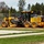 A John Deere 670GP motor grader is actively grading a dirt/gravel surface at what appears to be a construction or infrastructure development site. The machine is yellow and prominently branded with 'DEERE' and model number '670GP', with additional markings indicating '20-155' and a company name 'MATBY'. The site is bordered by a chain-link fence, stacked pipes or culverts, evergreen trees, and a paved area with grass in the foreground.