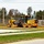 A yellow John Deere 670G motor grader is operating on a dirt/gravel surface near a paved road, likely performing grading or road preparation work. The scene appears to be a rural or semi-rural construction site with a fenced storage yard visible in the background. A red fire hydrant and utility poles are visible in the foreground area.