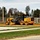 A yellow John Deere 670G motor grader is actively working on a construction or grading site, moving soil on what appears to be a newly developed area adjacent to a road. The machine is operating on reddish-brown disturbed earth, suggesting land preparation or road base grading work. A red fire hydrant is visible in the foreground near the roadside, indicating proximity to an established infrastructure zone.