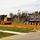 A road construction scene showing active grading and earthwork operations on what appears to be a new road or parking area. A yellow John Deere motor grader and a purple dump truck with a gray bed are working on freshly graded orange/red soil. A 'Road Closed Ahead' warning sign is prominently displayed in the foreground to alert drivers.