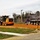 A road construction scene showing active earthwork operations on what appears to be a cleared and graded area with red/orange clay soil. A John Deere motor grader and a large dump truck are visible working on the site. A prominent orange 'Road Closed Ahead' warning sign is posted at the foreground right.