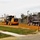 A road construction scene showing active grading and earthwork operations on what appears to be a road or parking area. A John Deere motor grader is working alongside a dump truck on freshly laid orange/red gravel or clay fill material. A 'Road Closed Ahead' warning sign is prominently displayed in the foreground.