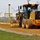 A yellow John Deere motor grader is actively grading a dirt/gravel road surface at a construction site. The machine is equipped with GPS/machine control masts on both sides, indicating precision grading technology is in use. The scene appears to be at or near a fenced facility with white buildings visible in the background.