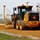 A yellow John Deere motor grader is actively grading a dirt/gravel road surface at a construction site. The machine is equipped with GPS mast receivers on both sides, indicating use of machine control technology for precision grading. The scene is set in a rural or semi-industrial area with utility poles, a chain-link fence, and commercial buildings visible in the background.