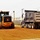 A road construction site showing a motor grader and a dump truck operating on freshly graded red clay/dirt surface. The grader appears to be leveling and smoothing the subgrade material that has likely been deposited by the dump truck. The scene is set in a rural or semi-rural area with a building and trees visible in the background.