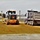 A road construction scene showing a yellow motor grader working alongside a dump truck on a freshly graded gravel or sand surface. The grader appears to be leveling and spreading material that has been deposited by the dump truck. The scene is set in what appears to be a small town or facility with white buildings visible in the background.
