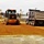 A road construction scene showing an orange motor grader actively grading a red clay/gravel surface alongside a large dump truck. The work appears to be taking place in a small town or institutional campus setting with white buildings visible in the background. The grader is equipped with GPS/laser guidance masts, indicating modern precision grading operations.