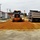 A road construction scene showing an orange motor grader spreading reddish-orange fill material (likely clay or gravel base) on a road surface. A dump truck is positioned ahead of the grader, likely having just deposited the fill material. The work is taking place in what appears to be a small institutional or military-style campus with white and red-roofed buildings on either side.