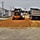 A road construction scene showing a yellow motor grader spreading and leveling reddish-brown gravel or crushed aggregate material on a roadway surface. A dump truck is positioned to the right, likely having recently deposited the material. The work is taking place in what appears to be a military installation or institutional campus with white buildings and red roofs visible in the background.