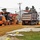 A road construction scene showing a John Deere motor grader working alongside a dump truck on a graded dirt surface. The work appears to be in a residential or institutional area with white and red-roofed buildings visible in the background. The grader is spreading and leveling reddish-brown gravel or fill material across what appears to be a new road base.
