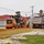 A road construction or grading operation is underway in what appears to be a residential or institutional campus setting. A John Deere motor grader in yellow-orange livery is working alongside a dump truck, spreading and leveling gravel or aggregate material. The scene suggests active site preparation, likely for a new road or parking area.