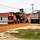 A road construction or grading operation is underway near a building with a red roof. A John Deere motor grader is actively grading a dirt/gravel surface alongside a large dump truck. The scene appears to be a parking lot or road base preparation project at what looks like a commercial or institutional facility.