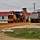 A construction site showing a John Deere motor grader working alongside a large dump truck on a graded dirt surface. The scene appears to be road or parking lot construction near white buildings with red roofs. The overcast sky and disturbed earth suggest active grading and material spreading operations.