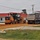 A road construction scene showing a John Deere motor grader working alongside a dump truck on a graded dirt surface. The work is taking place adjacent to a building complex with red roofs, likely a military installation or institutional campus. Orange safety cones and survey equipment are visible, indicating active grading and road preparation work.