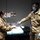 Two U.S. Army soldiers in OCP (Operational Camouflage Pattern) uniforms are engaged in what appears to be a medical or sanitation procedure, both wearing PPE including surgical masks, safety goggles, and nitrile gloves. One soldier is handing or spraying something into a clear plastic bag held by the other soldier at a table covered with a blue pad and paper towels. A posted notice on the wall behind them references N95 mask re-use protocols, suggesting this is related to COVID-19 PPE decontamination procedures.