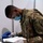 A U.S. Army soldier wearing OCP (Operational Camouflage Pattern) uniform is bent over a table, reviewing paperwork while handling medical or testing equipment. The soldier is wearing personal protective equipment including a blue surgical mask and clear safety goggles, along with latex gloves. The setting appears to be a temporary or field medical/testing station.
