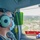 A military or aviation crew member wearing aviation headsets, safety glasses, and a face mask looks out the window of an aircraft in flight. The aerial view below shows a suburban or semi-urban landscape with roads, fields, and structures. A folded American flag is visible in the lower right corner of the frame, suggesting a ceremonial or memorial mission.