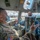 Two military personnel wearing face masks are seated in the cockpit of a large military aircraft, appearing to be engaged in a briefing or discussion. The individual in the foreground wears U.S. Army camouflage (OCP pattern) with an American flag patch, while the person in the background appears to be an aircrew member. The setting appears to be on a flight line at a military airfield, visible through the cockpit windows.