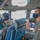 Two military aircrew members are seated in the cockpit of a large military aircraft, likely a C-17 Globemaster or similar transport aircraft. Both personnel are wearing olive drab flight suits with U.S. flag patches and face masks, suggesting the photo was taken during the COVID-19 pandemic era. An American flag is visible folded in the background between the crew members.