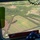 An aerial view photographed from inside a military or government aircraft, looking down over a green, hilly landscape with agricultural fields and wind turbines. A folded American flag is visible in the foreground on what appears to be a wooden surface inside the aircraft. The terrain below shows a mix of farmland, forested areas, and a road, consistent with a Hawaiian landscape.