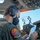 A military aviator wearing a Bose headset and black face mask is seated in what appears to be a large military transport aircraft cockpit, reviewing a navigation chart or approach plate. The pilot is wearing an olive drab flight suit with a squadron patch visible on the shoulder reading '335th Airlift Squadron.' The cockpit displays numerous avionics panels, illuminated indicators, and digital navigation screens.