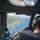 A pilot is seated in the cockpit of an aircraft flying over a tropical coastline, likely Hawaii, based on the lush green terrain and turquoise waters visible below. The view through the large cockpit windows shows a sweeping aerial perspective of a curved shoreline with white surf, green agricultural fields, and distant cliffs. The pilot is wearing aviation headphones and the cockpit displays modern avionics equipment.