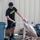 A young man wearing a black U.S. Army physical training t-shirt is using a water hose to spray the inside of a large brown paper bag outdoors. He appears to be cleaning or rinsing the bag near a metal building wall. Several black trash bags and debris are visible in the background.