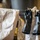 Two black rubber industrial gloves are hanging on a clothesline or wire, secured with wooden clothespins, alongside white protective garments or Tyvek suits. The gloves appear heavily soiled with a dark, oily or chemical substance. The setting suggests a work or industrial environment where protective equipment is being dried after use.