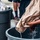 A person is wringing out or submerging a wet tan/beige fabric bag or garment into a large dark plastic barrel or tub filled with water. Water is visibly dripping from the fabric. Multiple large dark plastic containers are present, and several people are partially visible in the background.