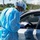 A healthcare worker in full personal protective equipment (PPE) is leaning toward the open window of a silver car, holding a blue clipboard. The setting appears to be an outdoor drive-through testing or screening site. The worker is wearing a disposable gown, surgical mask, face shield, hair cover, and latex gloves.