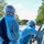 Two healthcare workers in full personal protective equipment (PPE) are conducting a drive-through medical procedure, likely COVID-19 testing, at an outdoor site. Both workers are wearing blue isolation gowns, surgical masks, hair covers, and protective eyewear. One worker is leaning toward the open window of a silver vehicle while the other stands nearby observing.