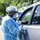 A healthcare worker in full personal protective equipment (PPE) is conducting a drive-through testing or vaccination service beside a silver vehicle. The worker is wearing a blue disposable gown, hair cap, face mask, face shield, and latex gloves, and appears to be handling a small vial or specimen container. The setting appears to be an outdoor parking lot during daytime with green trees visible in the background.