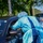 The image depicts a drive-through medical testing or screening operation, likely a COVID-19 testing site. Two healthcare workers in full personal protective equipment (PPE) are attending to a person seated in a dark-colored vehicle. A third individual in military camouflage uniform is partially visible on the right edge of the frame.