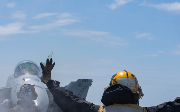 A U.S. Navy flight deck crew member designated 'FLY 3' signals with both arms raised toward an F/A-18 fighter jet on an aircraft carrier flight deck. Steam or exhaust is visible around the aircraft, suggesting catapult launch preparation. Additional crew members in red and yellow gear are visible in the background.