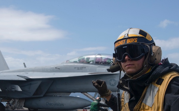 A U.S. Navy flight deck crew member stands on an aircraft carrier flight deck wearing a yellow safety vest and helmet marked 'BUCKET BOYS', giving hand signals. Behind them is a U.S. Navy F/A-18 Hornet jet fighter with 'CheckMates' written on its fuel tank, belonging to squadron VFA-211. The scene depicts standard carrier flight deck operations at sea.