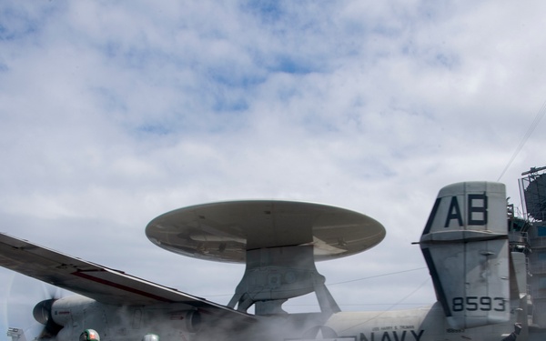 A U.S. Navy E-2 Hawkeye airborne early warning aircraft (bureau number 8593, tail code AB) is being launched from an aircraft carrier flight deck. Steam from the catapult system is visible as flight deck crew in green jerseys crouch and signal during the launch sequence. Additional aircraft and the carrier's island superstructure are visible in the background.