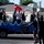 A parade float decorated with a blue wave design carries several people holding umbrellas of various colors. The float displays a sign reading 'TXT3 Weather the Storm under our Umbrella'. Spectators on the street are waving and interacting with the float participants.
