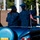 Two graduates in navy blue caps and gowns are seated in the back of a classic Chevrolet convertible, likely participating in a graduation parade or procession. The vehicle displays two small American flags and bears a yellow license plate reading '2537 HC'. One graduate is waving to the crowd while both face away from the camera.