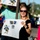 A smiling woman wearing sunglasses and a black top holds a handmade sign reading 'YOU DID IT!' featuring the HHS (likely a high school) mascot, a yellow jacket/hornet. The scene appears to be an outdoor celebration, possibly a graduation parade or similar school event. Other people are visible around her, also holding signs and cheering.
