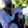 The image shows a group of graduates at an outdoor graduation ceremony, all wearing face masks along with their caps and gowns. At least three graduates are visible, wearing different colored gowns (white, blue, and dark red/maroon), suggesting they may be from different schools or honor groups. The presence of face masks indicates this ceremony took place during the COVID-19 pandemic period.